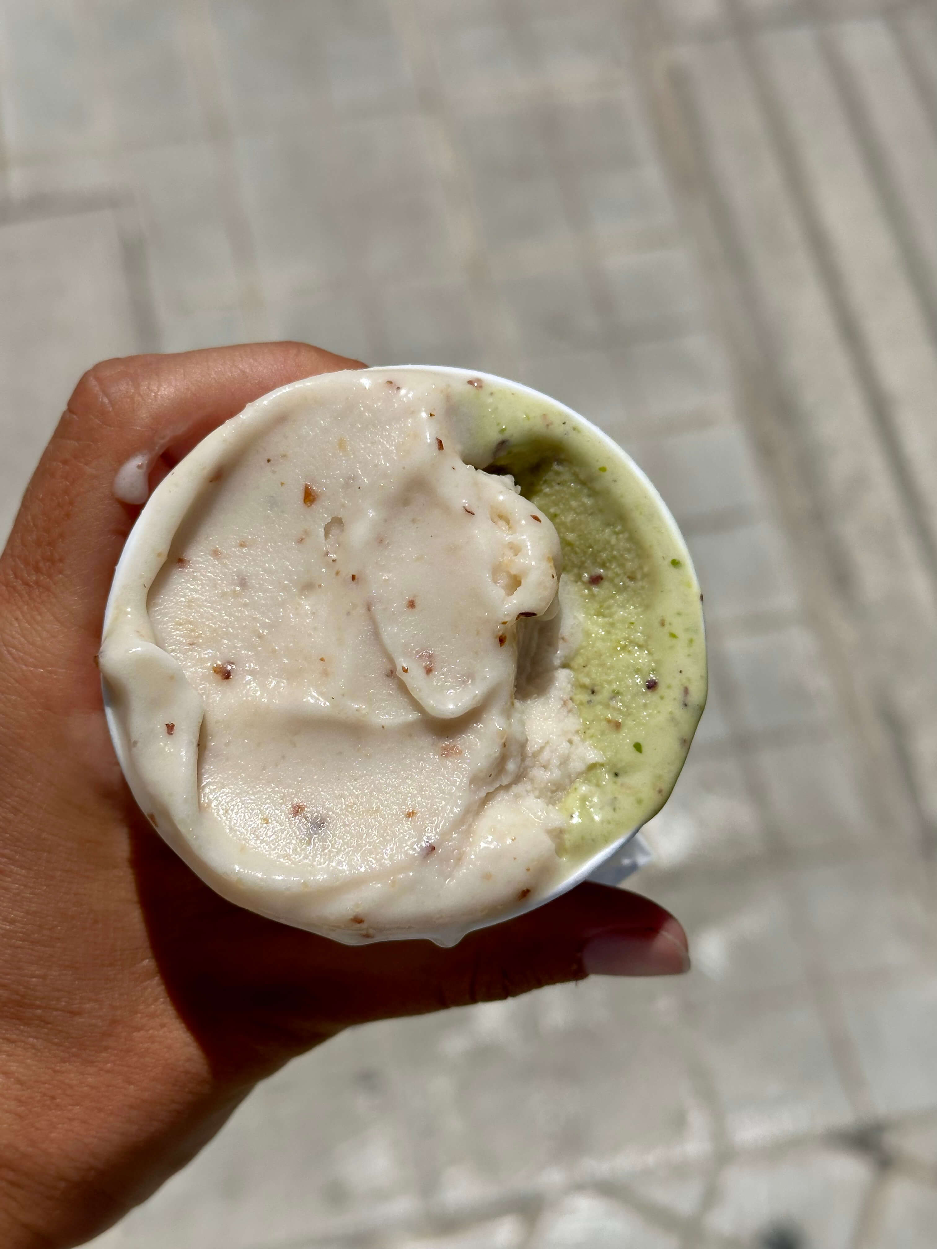 A hand holds a cup of two-flavor gelato: light-colored with specks and green pistachio.