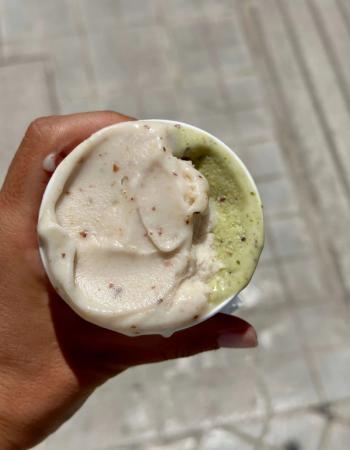 A hand holds a cup of two-flavor gelato: light-colored with specks and green pistachio.
