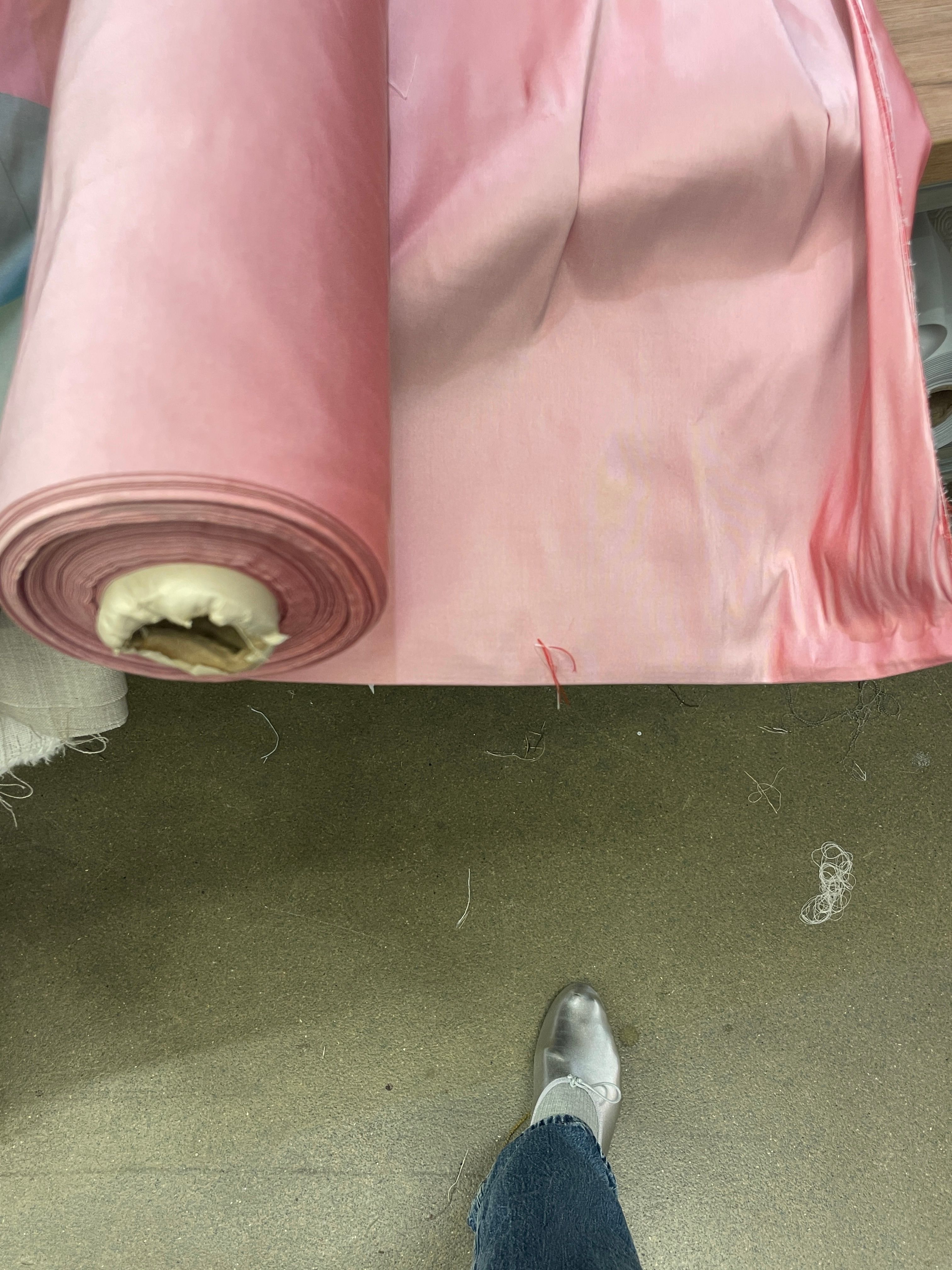 A large roll of light pink fabric unwinds onto a grey floor, with a person's foot in a silver shoe visible.