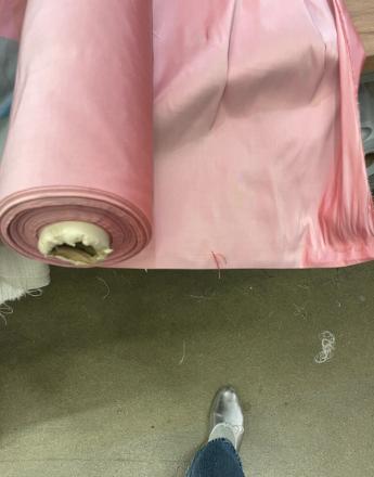 A large roll of light pink fabric unwinds onto a grey floor, with a person's foot in a silver shoe visible.