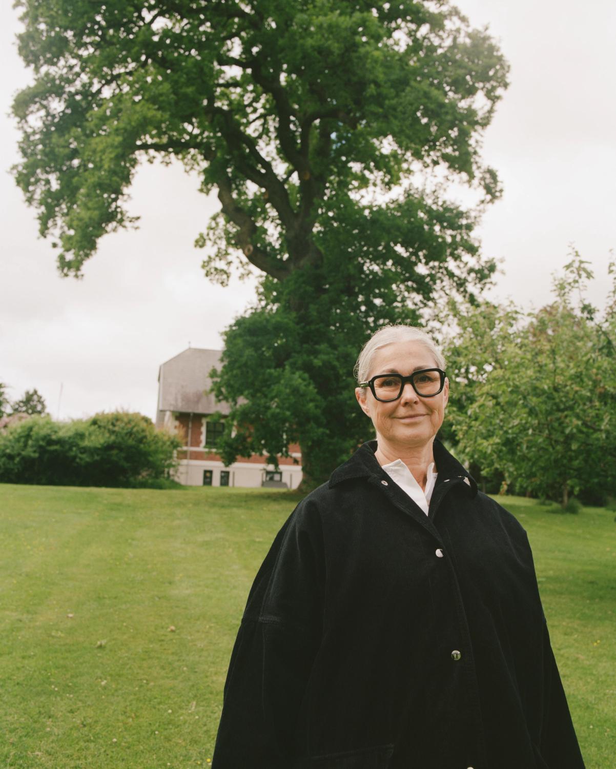 A woman with grey hair, black-framed glasses, and a black oversized coat smiles faintly outdoors with a large tree and building in the background.