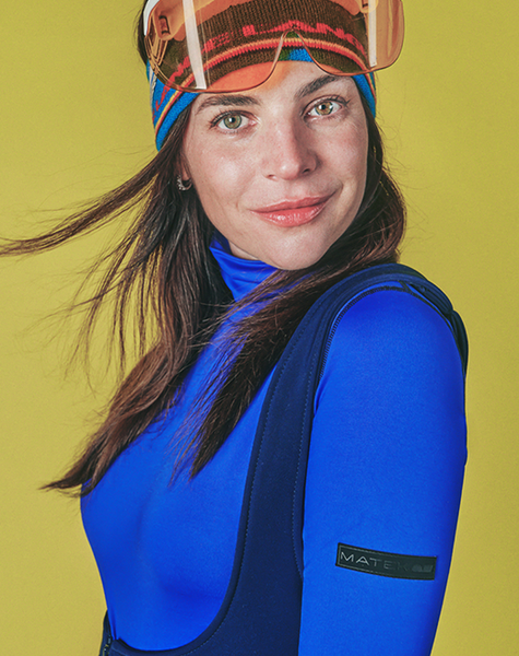 A woman with ski goggles pushed up, holding a blue snowboard with a pink R logo, against a yellow background.