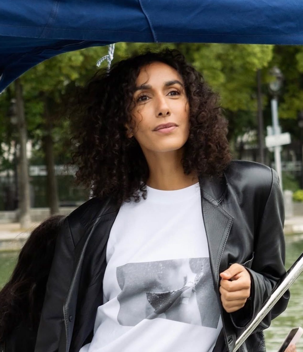 A woman with dark, curly hair in a black leather jacket and white t-shirt looks thoughtfully from a boat under a blue canopy.