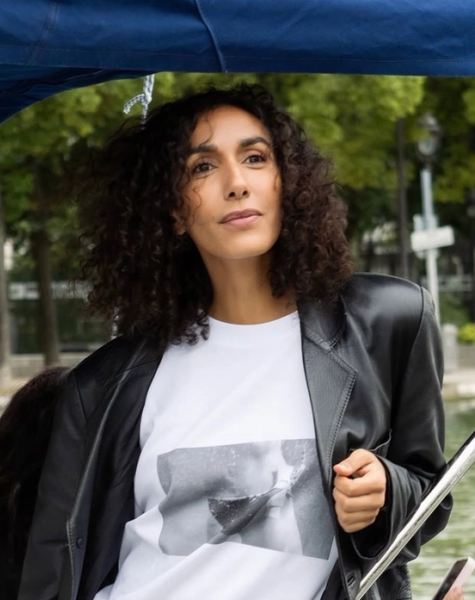 A woman with dark, curly hair in a black leather jacket and white t-shirt looks thoughtfully from a boat under a blue canopy.