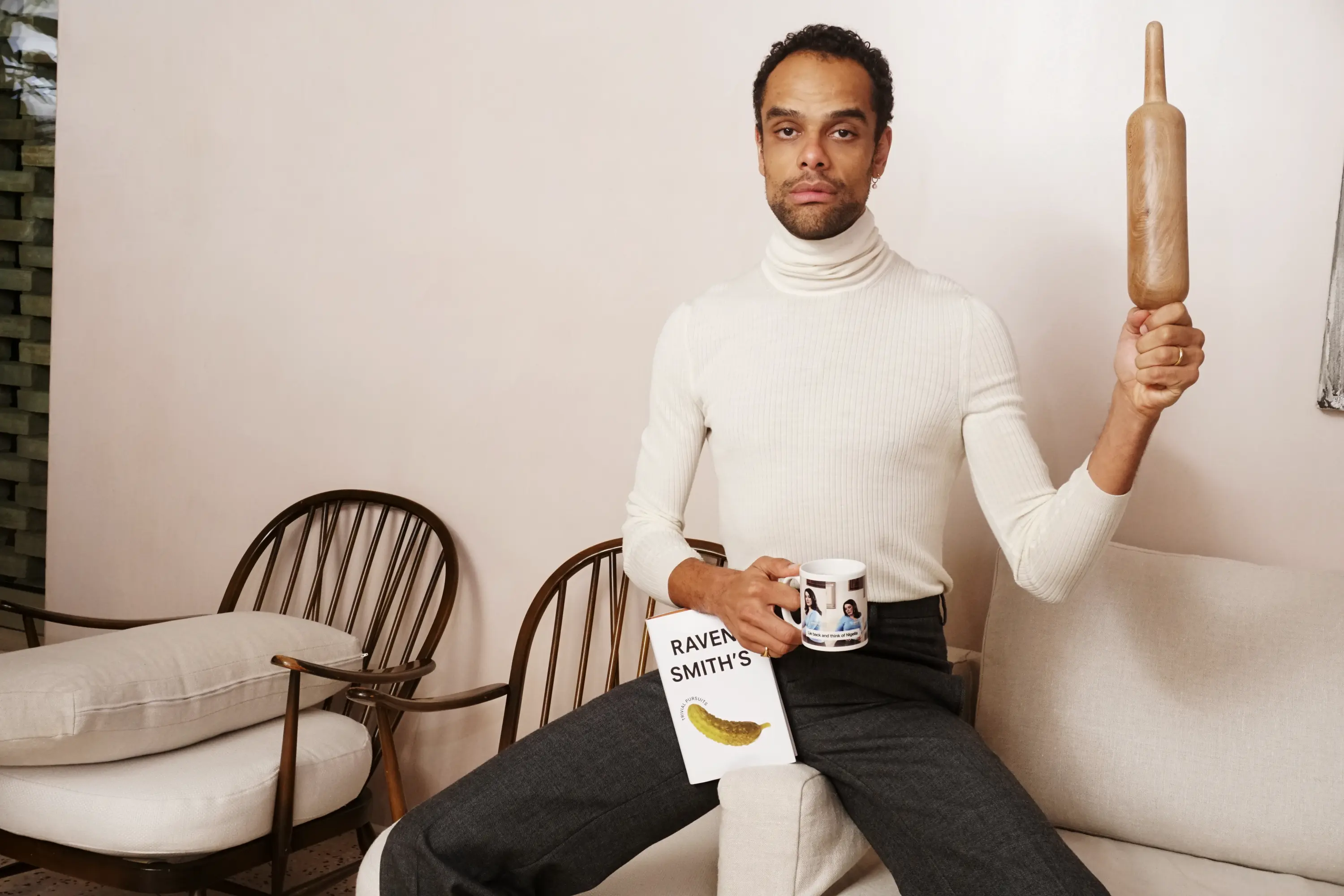 A man in a white turtleneck holds a book with a pickle on the cover, a mug depicting two women, and a wooden rolling pin held vertically, looking forward.