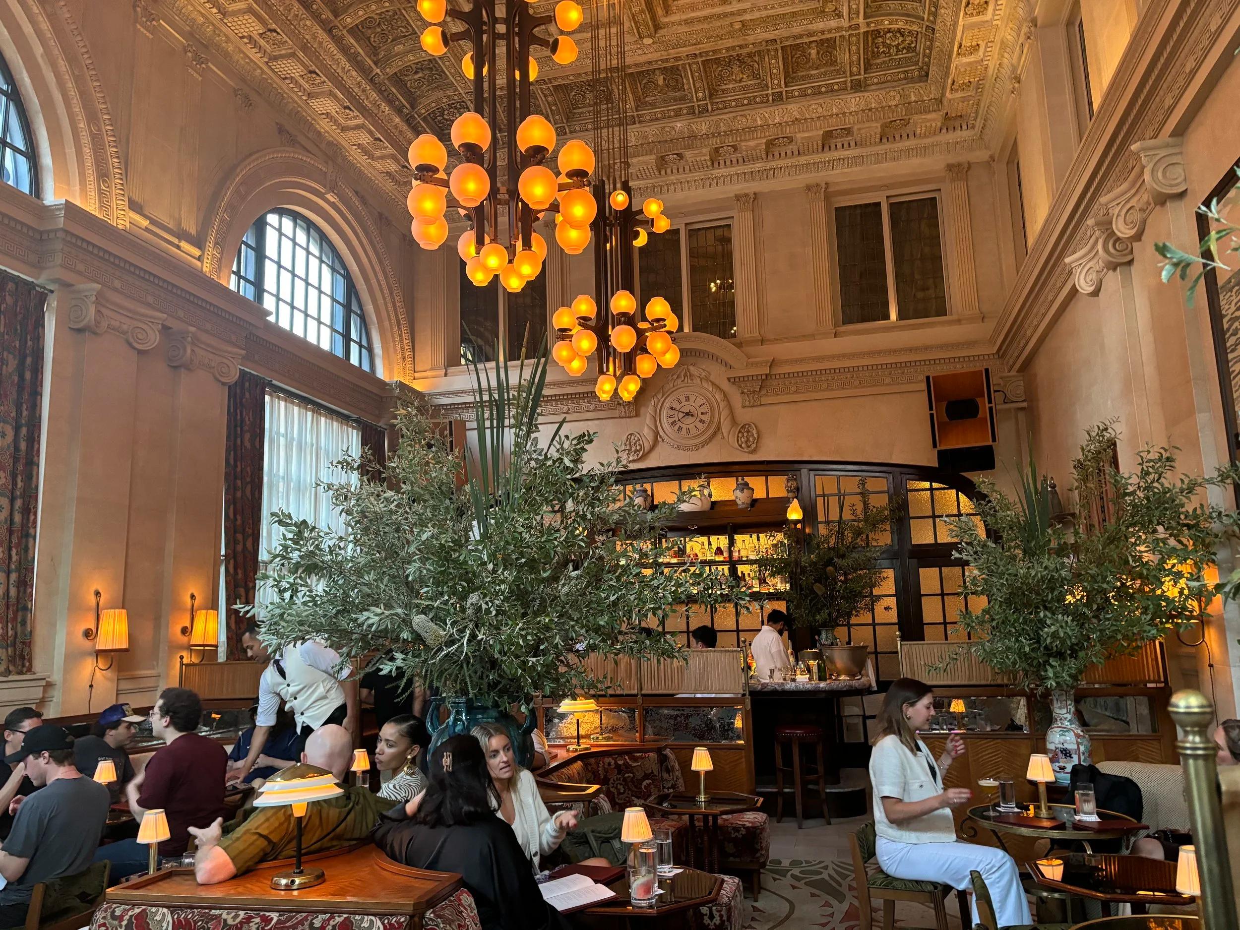 An opulent hotel lounge or bar with warm amber spherical chandeliers, lush potted plants, and many patrons socializing.