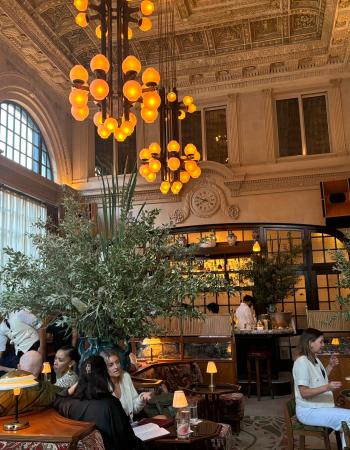 An opulent hotel lounge or bar with warm amber spherical chandeliers, lush potted plants, and many patrons socializing.