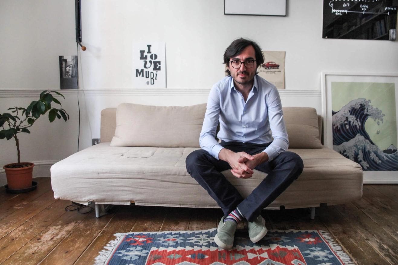 A man with glasses and a blue shirt sits cross-legged on a beige couch in a room with art prints, a plant, and a patterned rug on a wooden floor.
