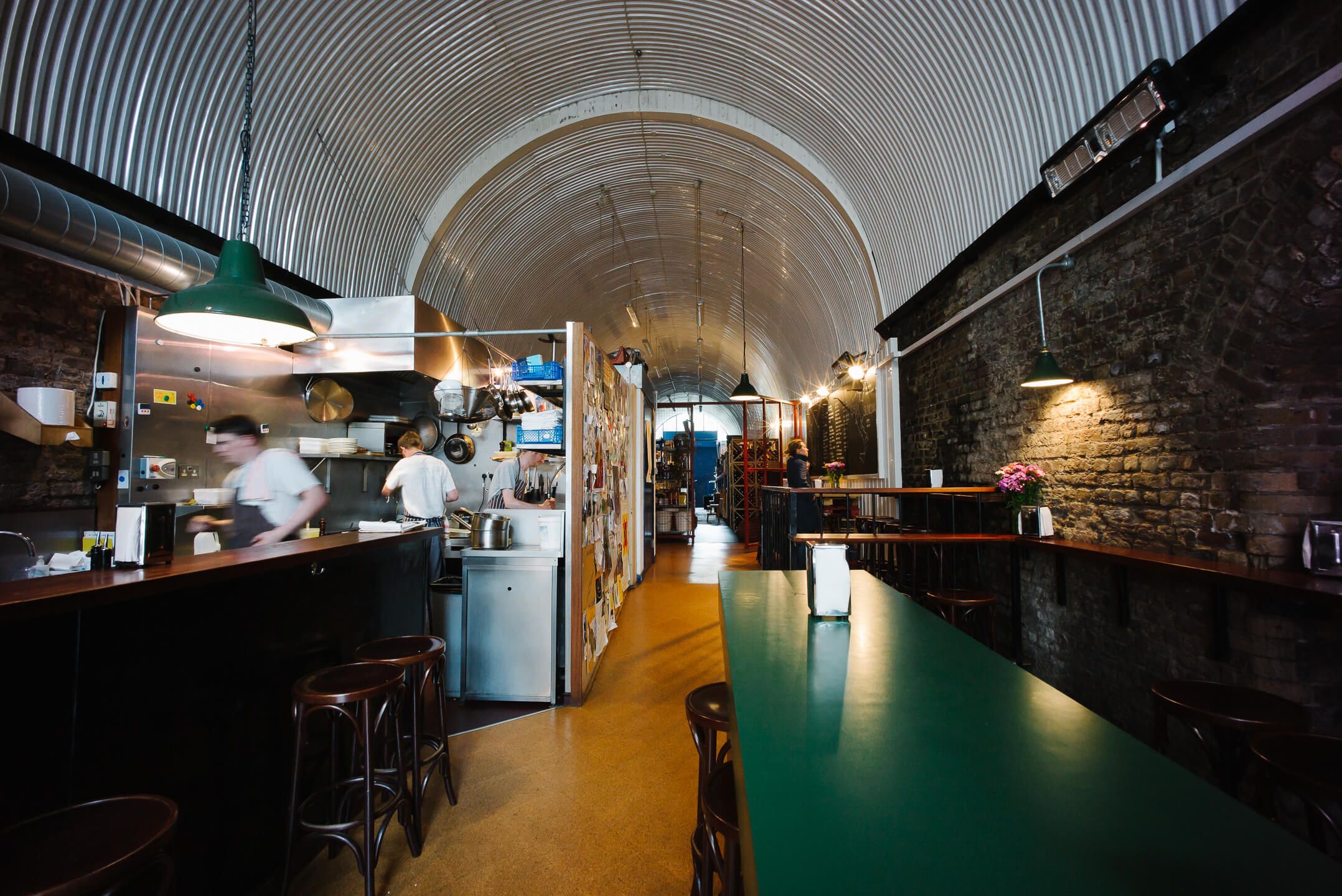 Interior of a long, narrow restaurant in an arched space with a corrugated metal ceiling, an open kitchen, exposed brick wall, and a green communal table.