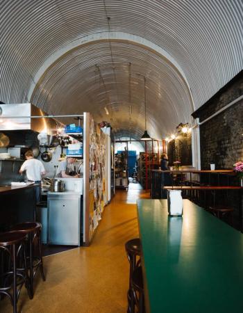 Interior of a long, narrow restaurant in an arched space with a corrugated metal ceiling, an open kitchen, exposed brick wall, and a green communal table.