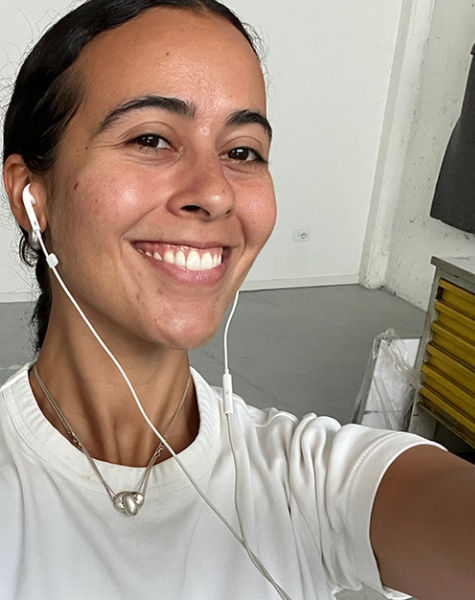 A smiling woman with dark hair pulled back takes a selfie, wearing a white t-shirt and wired earphones.