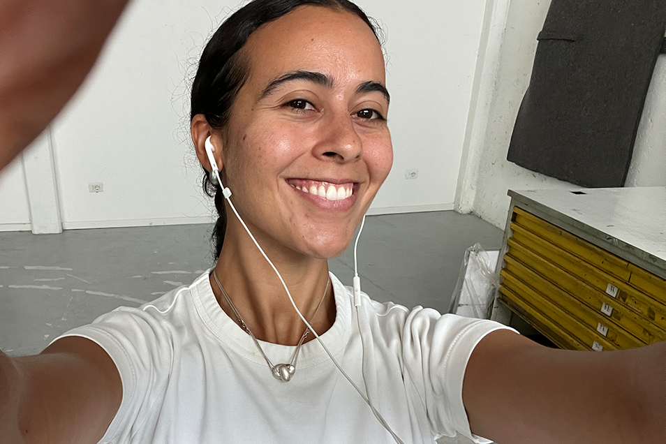 A smiling woman with dark hair pulled back takes a selfie, wearing a white t-shirt and wired earphones.
