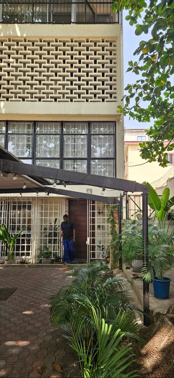 A man in a dark shirt and blue pants exits a building with a patterned wall, large windows, an awning, and many potted plants.