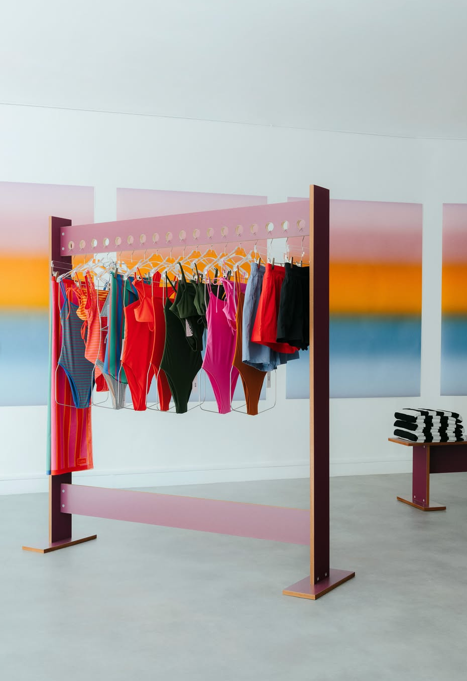 A modern pink clothing rack displays colorful swimsuits and garments against gradient art panels, with a stack of striped towels on a bench to the right.