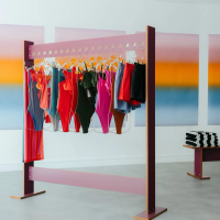 A modern pink clothing rack displays colorful swimsuits and garments against gradient art panels, with a stack of striped towels on a bench to the right.