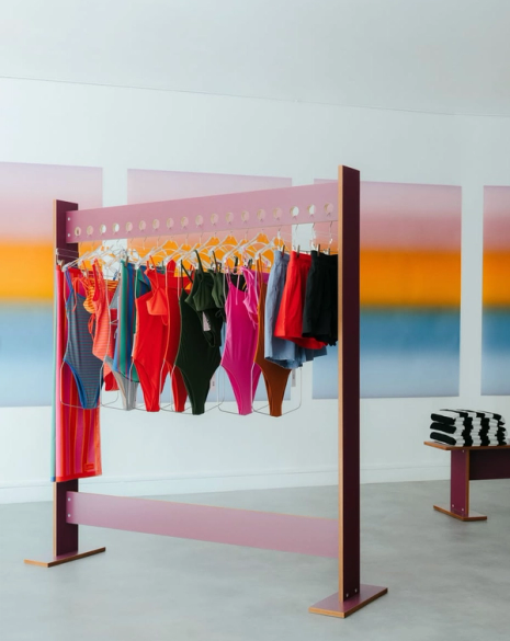 A modern pink clothing rack displays colorful swimsuits and garments against gradient art panels, with a stack of striped towels on a bench to the right.
