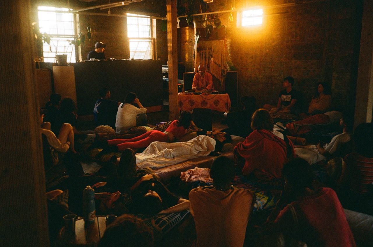 A dimly lit, rustic room with people gathered on mats, with a person at a music console on the left and a robed figure at a low table on the right.