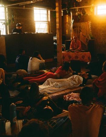 A dimly lit, rustic room with people gathered on mats, with a person at a music console on the left and a robed figure at a low table on the right.