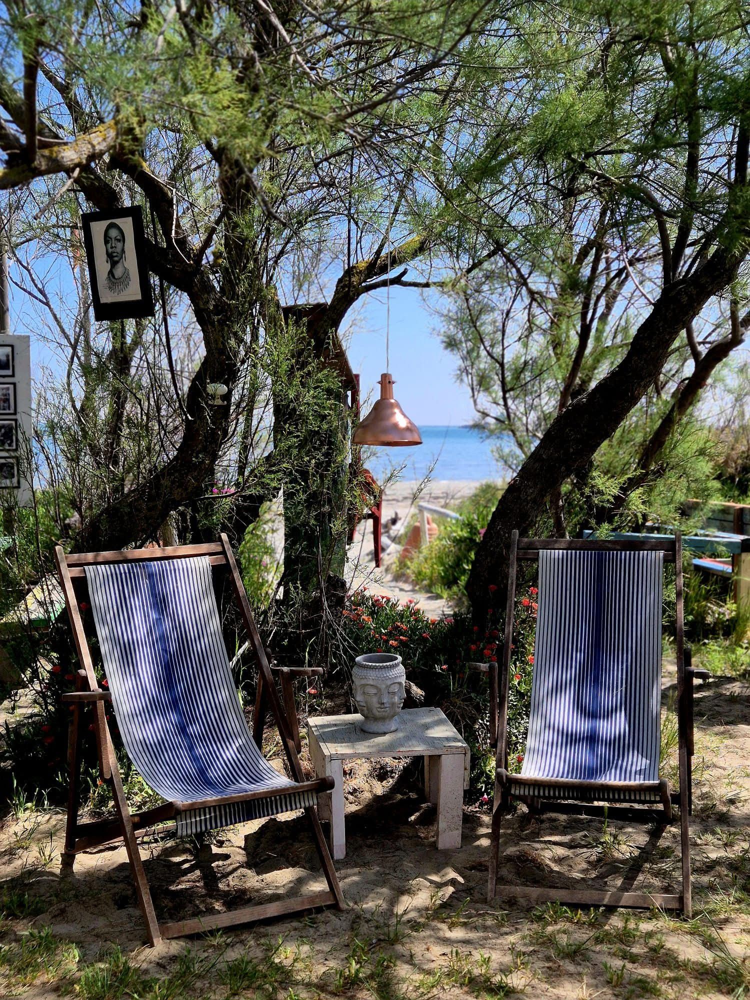 Two striped deck chairs and a table with a Buddha head sit under trees, overlooking a beach and sea. A portrait and copper lamp hang from the branches.