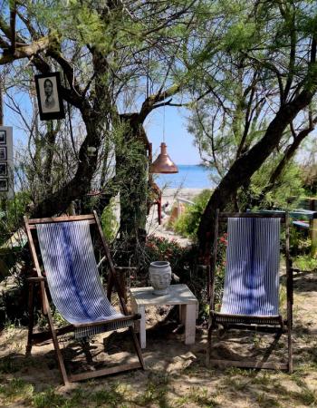 Two striped deck chairs and a table with a Buddha head sit under trees, overlooking a beach and sea. A portrait and copper lamp hang from the branches.