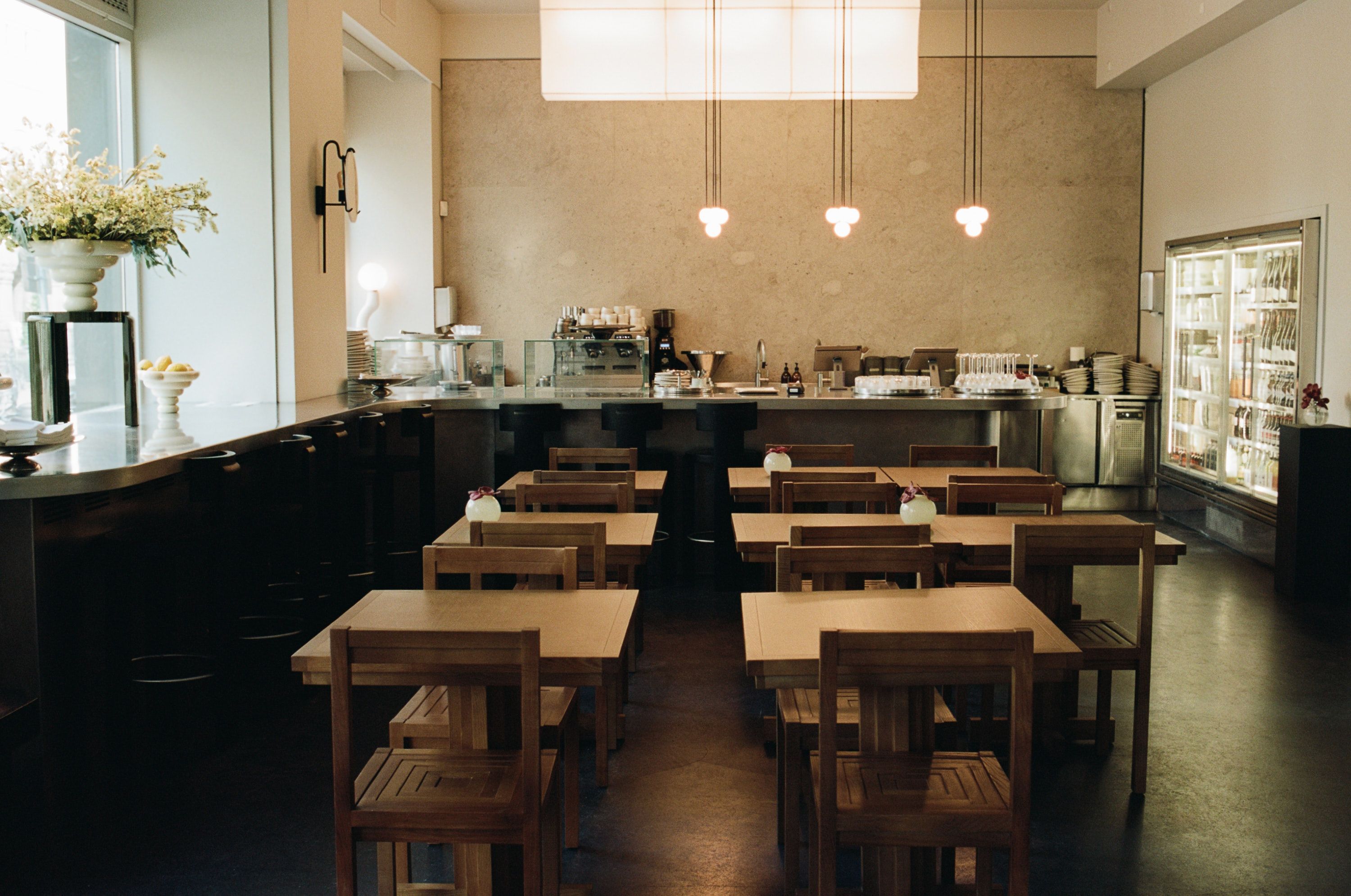 Interior of a clean, empty cafe with rows of wooden tables and chairs, a long counter, and a service area.