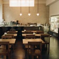 An empty, modern cafe interior with rows of wooden tables and chairs, a long bar, and a service counter with a glass refrigerator.