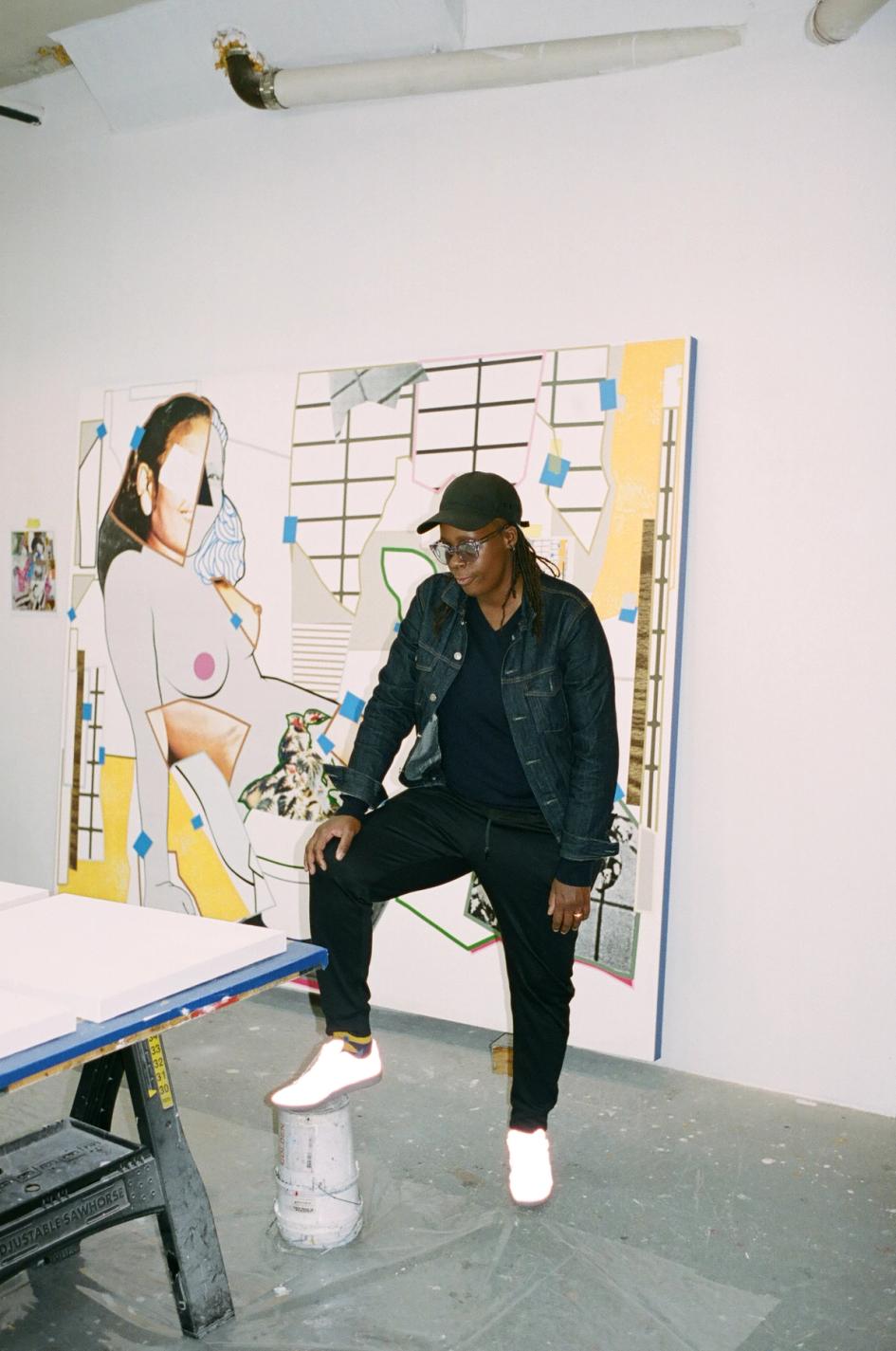 An artist in a denim jacket and cap stands with one foot on a paint bucket, in front of a large painting of a reclining female figure.