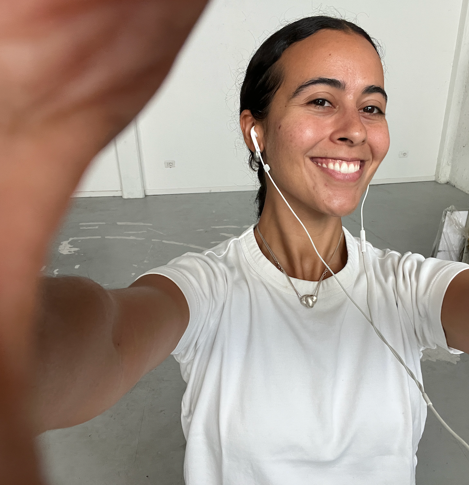 A smiling woman with dark hair pulled back takes a selfie, wearing a white t-shirt and wired earphones.