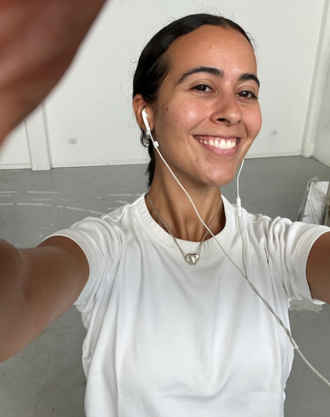 A smiling woman with dark hair pulled back takes a selfie, wearing a white t-shirt and wired earphones.