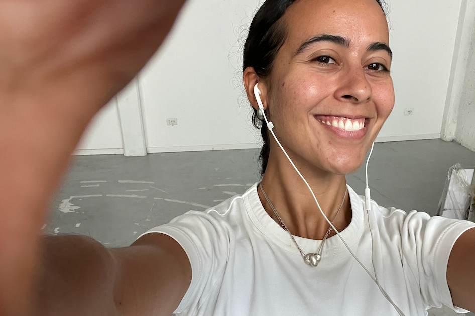A smiling woman with dark hair pulled back takes a selfie, wearing a white t-shirt and wired earphones.