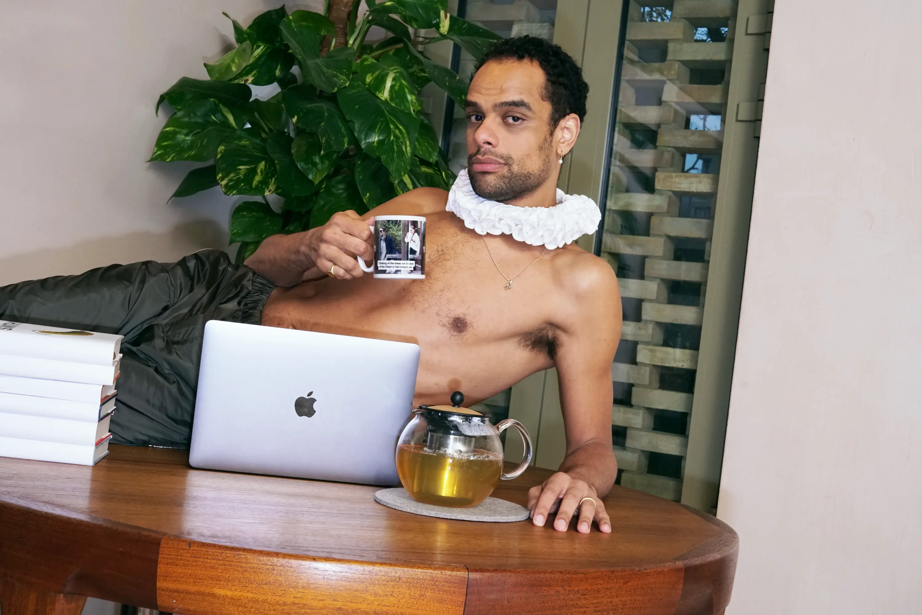 A bare-chested man with a beard and a white ruffled collar reclines, holding a mug, while looking at the camera. A laptop and a teapot of tea are on the table next to him.