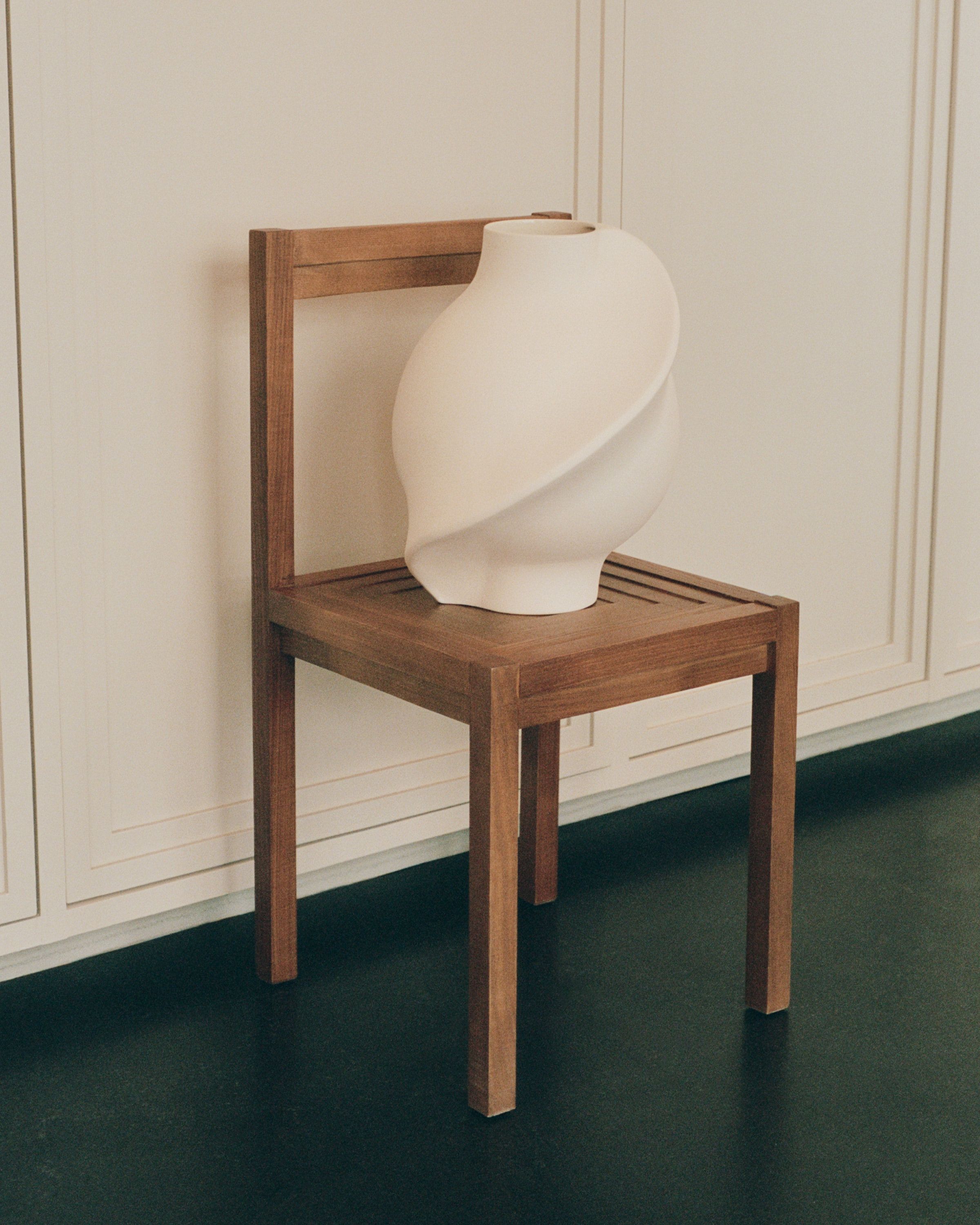A white, sculptural vase with a twisted design sits on a wooden chair.