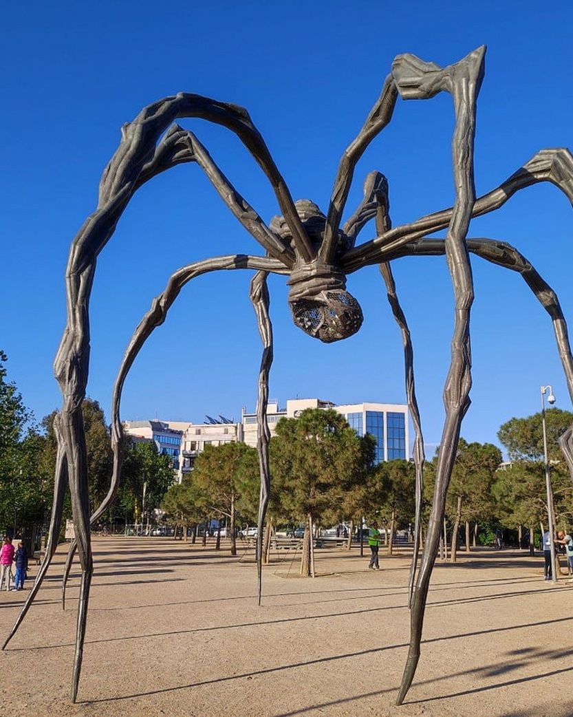 A massive bronze spider sculpture with an egg sac hangs underneath, standing in a park against a blue sky.