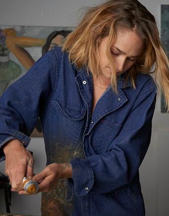 A woman in a black bra paints a female nude figure on an easel.