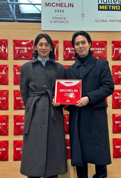 Two men in long coats holding a red Michelin 2026 award plaque.