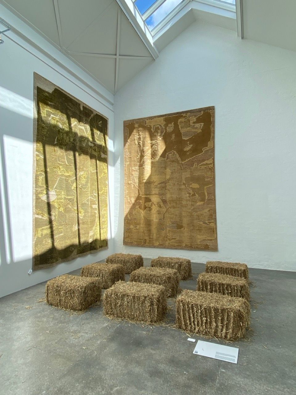 An exhibition featuring two large wall tapestries, one green and one brown, and nine hay bales arranged on a concrete floor under a skylight.