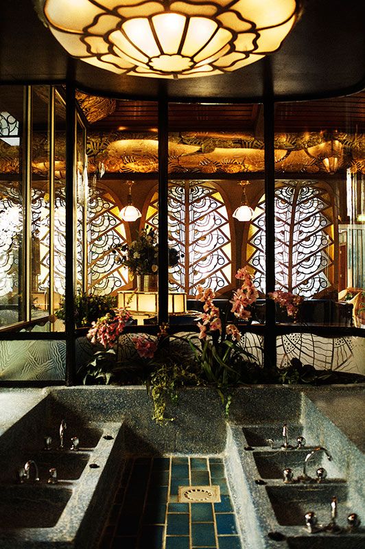 An ornate Art Deco interior featuring a large multi-basin stone sink, intricate geometric stained-glass windows, and a lotus-petal ceiling light.