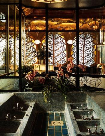 An ornate Art Deco interior featuring a large multi-basin stone sink, intricate geometric stained-glass windows, and a lotus-petal ceiling light.