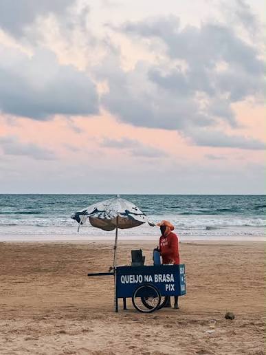 A vendor sells grilled cheese from a blue cart with an umbrella on a sandy beach, with the ocean and a pink-tinged sky in the background.