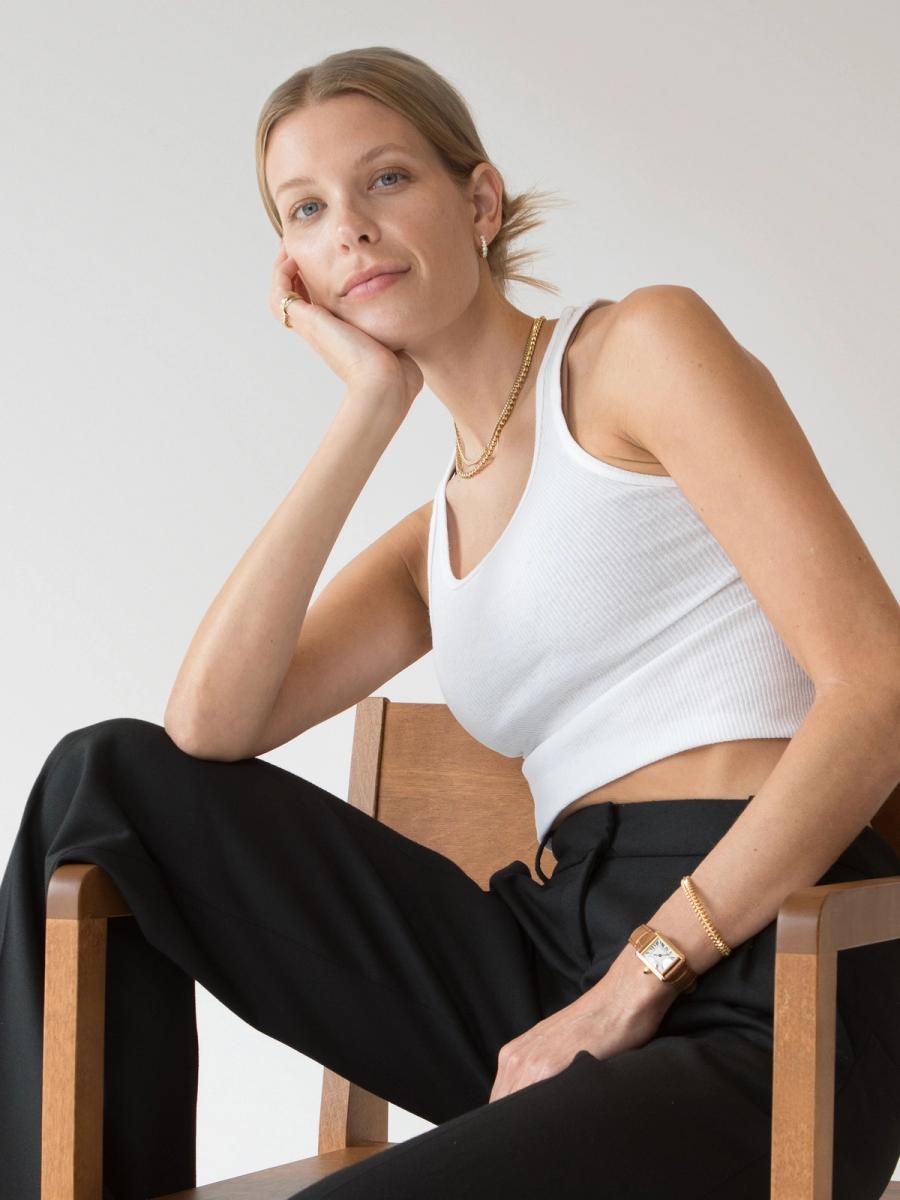 A woman in a white tank top and black pants sits on a wooden chair, resting her chin on her hand. She wears gold jewelry and a watch.