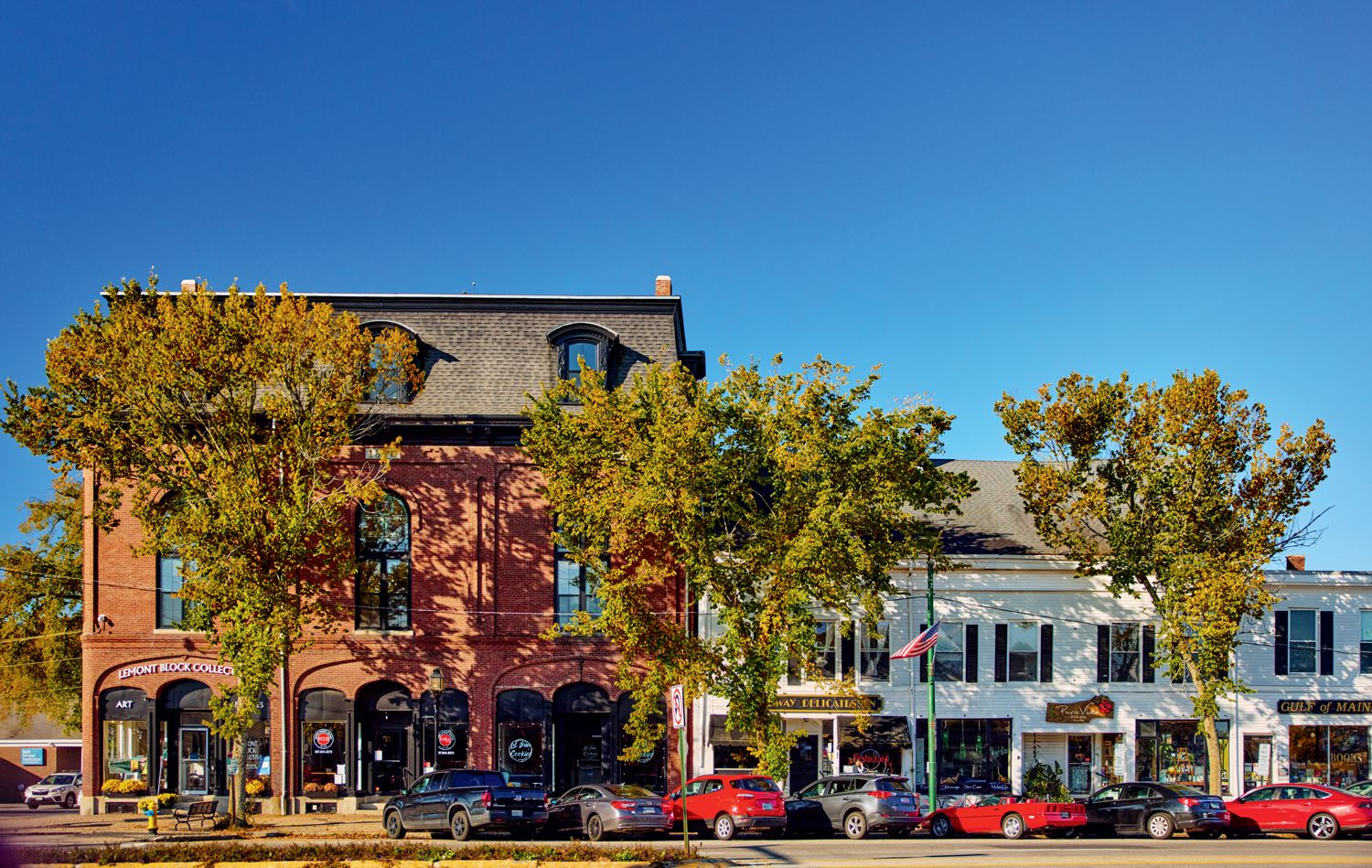 A street scene with brick and white buildings, yellow-green autumn trees, and parked cars under a clear blue sky.