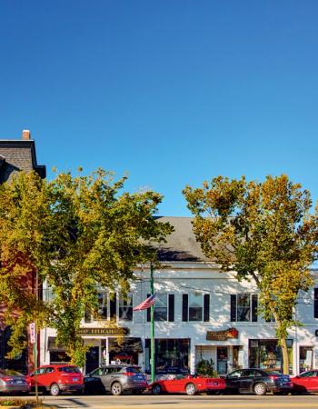 A street scene with brick and white buildings, yellow-green autumn trees, and parked cars under a clear blue sky.