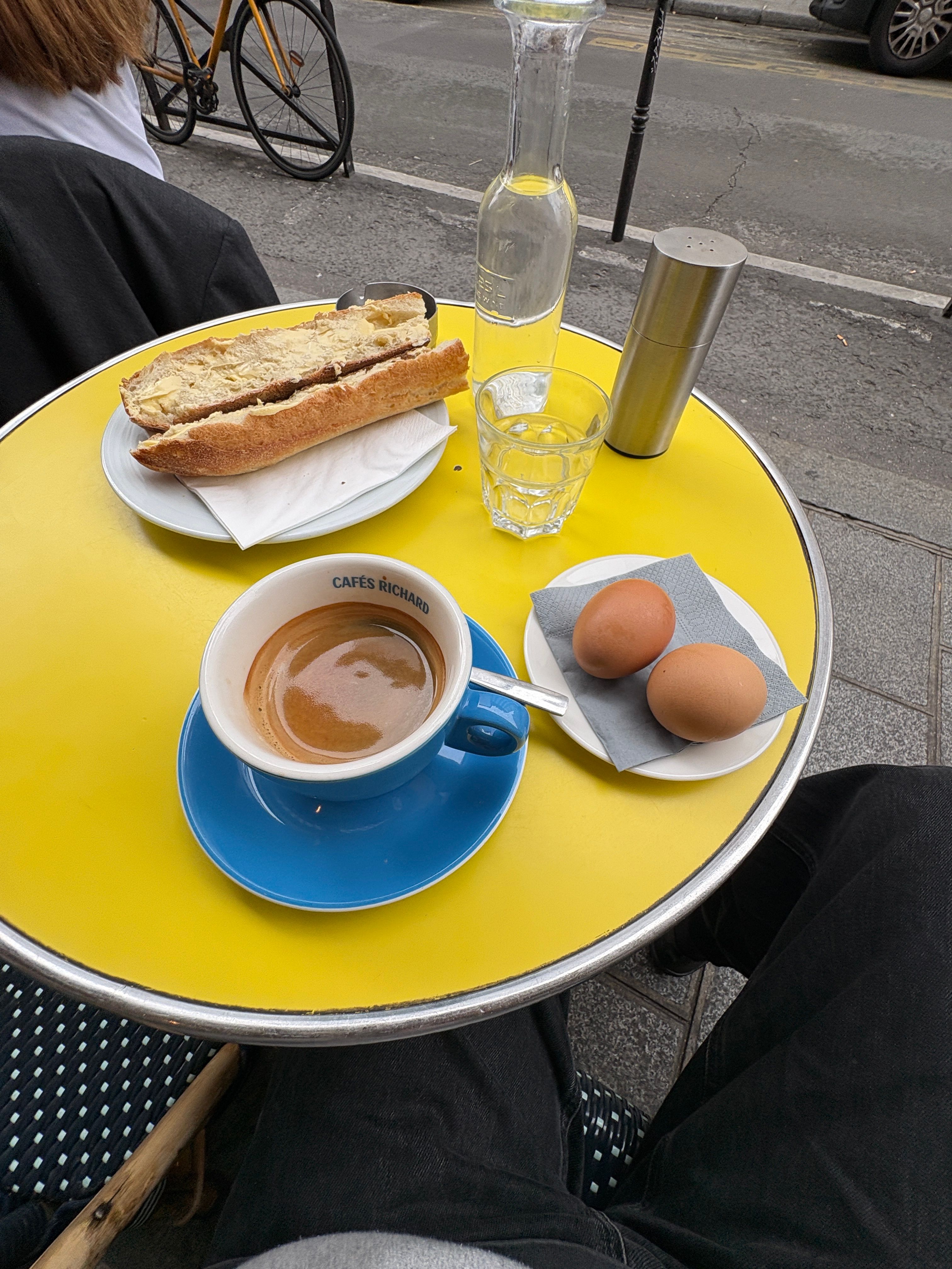 A yellow cafe table seen from above, holding a buttered baguette, coffee in a blue cup, two eggs, water, and a silver grinder. A person's legs are visible below, with a street in the background.