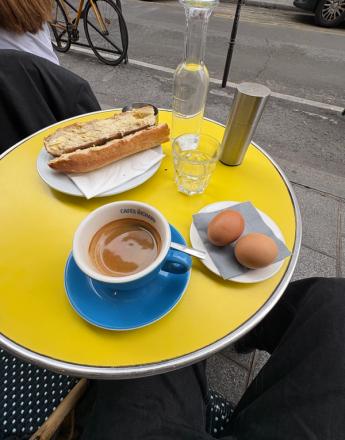 A yellow cafe table seen from above, holding a buttered baguette, coffee in a blue cup, two eggs, water, and a silver grinder. A person's legs are visible below, with a street in the background.