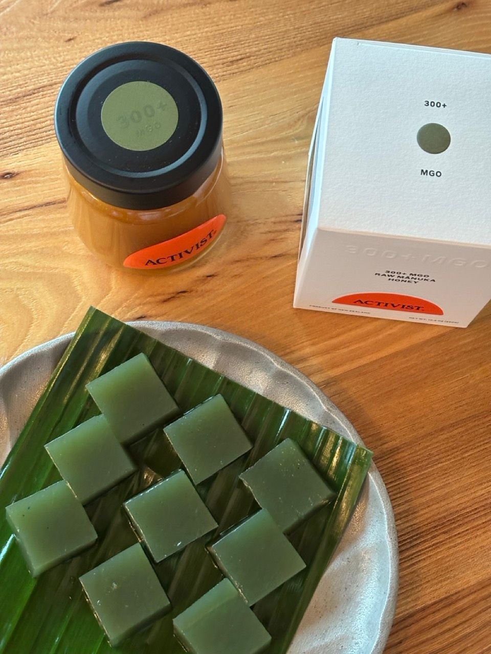 A jar of Activist 300+ MGO Raw Manuka Honey, its white box packaging, and green square jellies on a banana leaf on a plate.