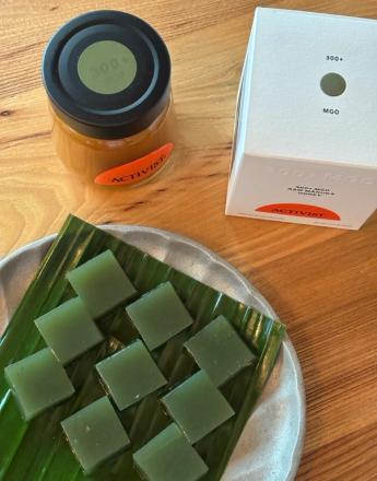 A jar of Activist 300+ MGO Raw Manuka Honey, its white box packaging, and green square jellies on a banana leaf on a plate.