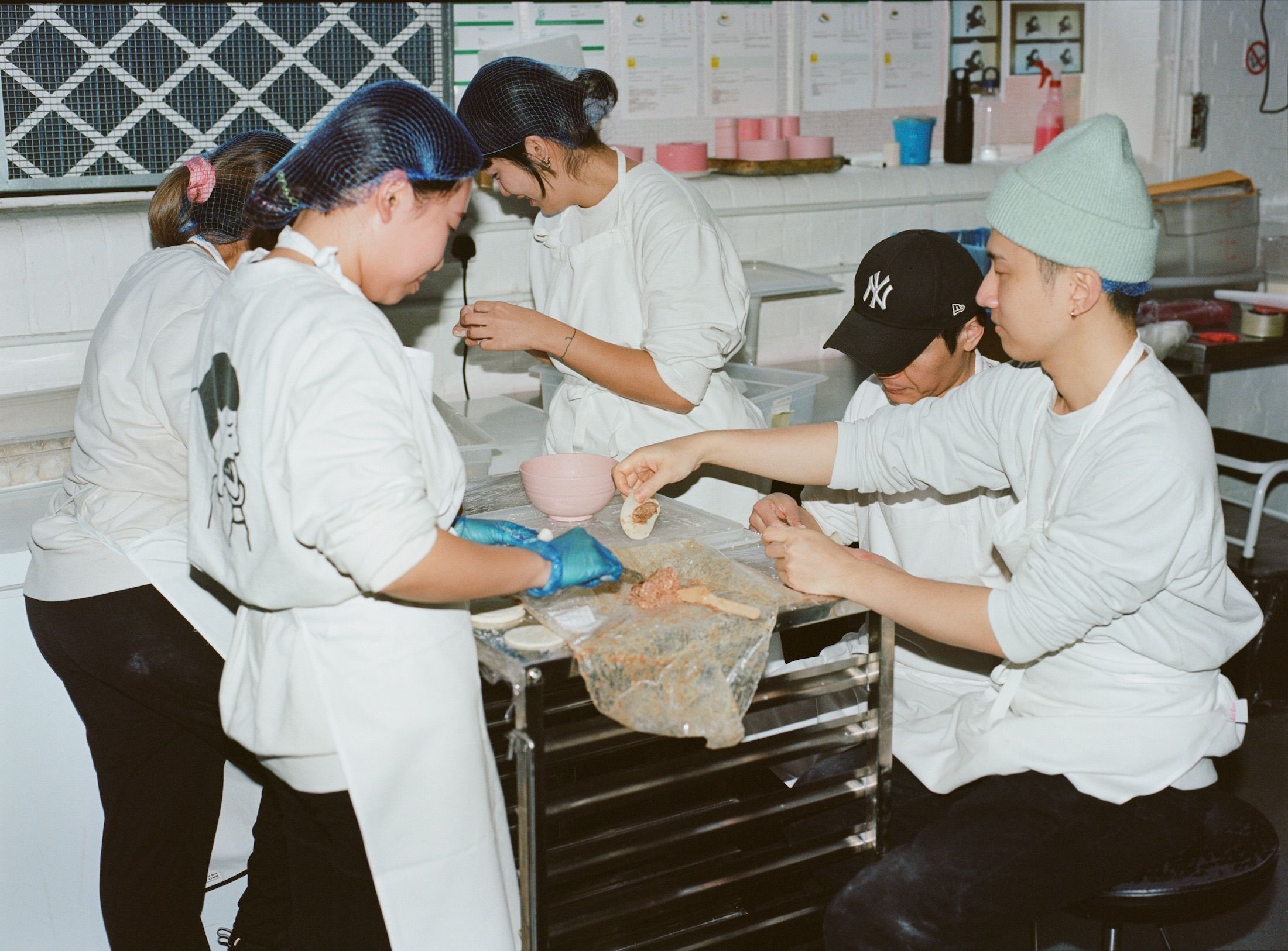 A group of people wearing aprons and hairnets assemble dumplings in a kitchen.