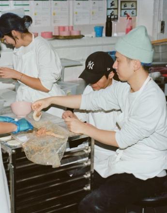 A group of people wearing aprons and hairnets assemble dumplings in a kitchen.