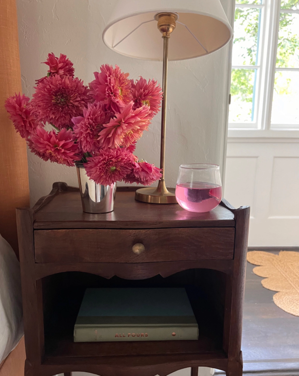A dark wooden nightstand holds a silver vase of pink dahlias, a brass lamp, and a glass of pink liquid; a book is on its lower shelf.