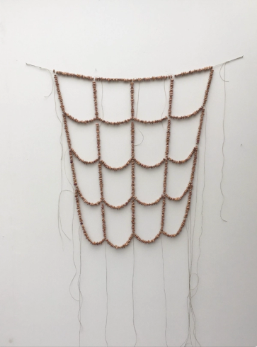 A net of small copper-colored beads forming a scalloped pattern, hanging on a white wall with loose threads.